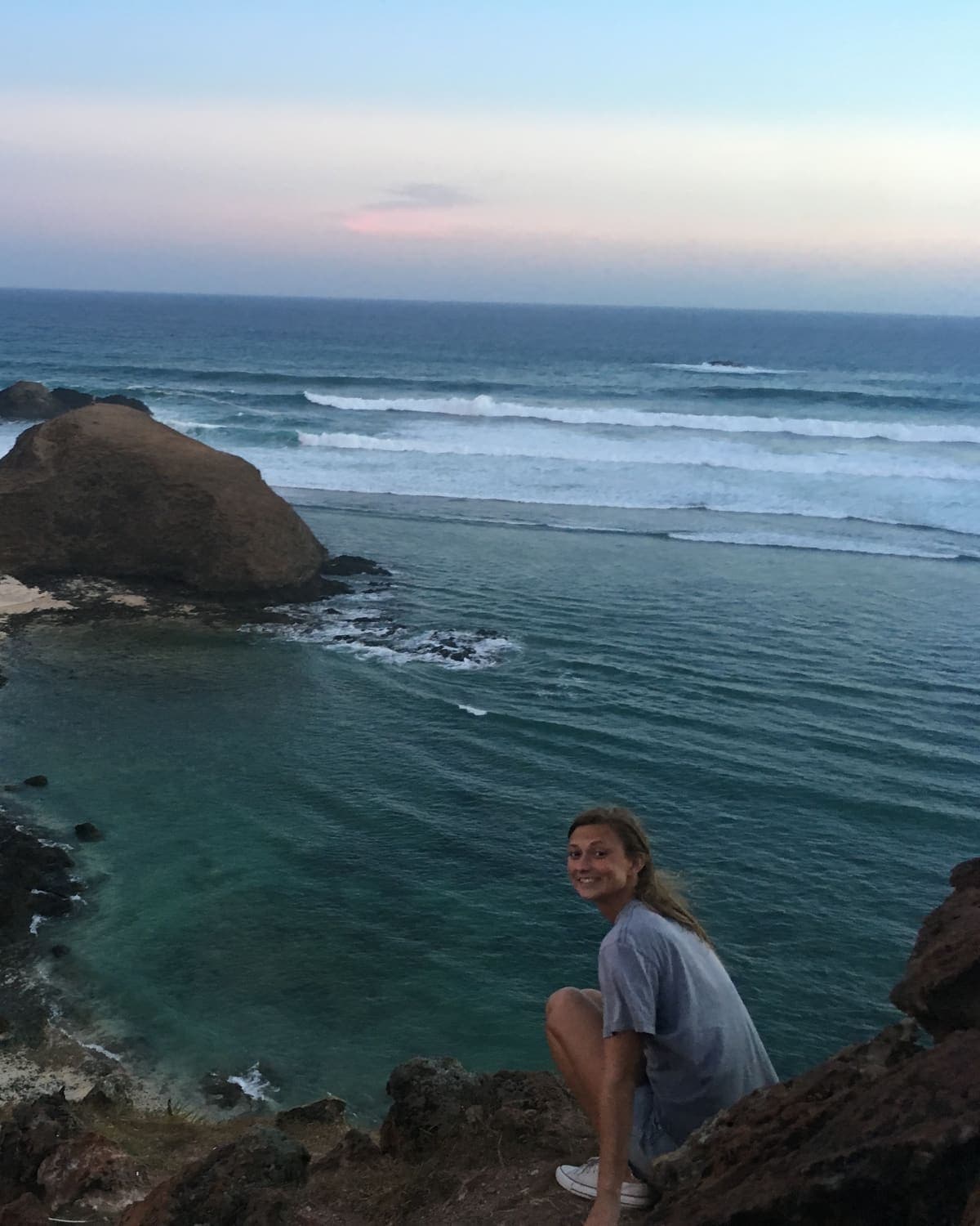 Ella crouched on rocks above a sheltered cove at dusk, smiling back over her shoulder, with lines of surf rolling in behind her.