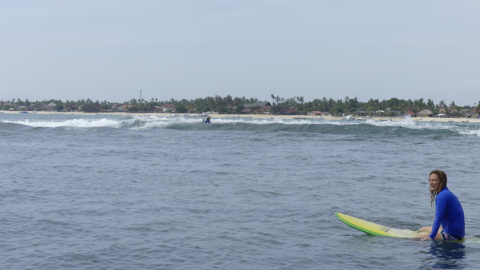 Ella sitting upright on a surfboard in calm blue water, smiling, with a hazy tree-lined shore in the distance behind her.