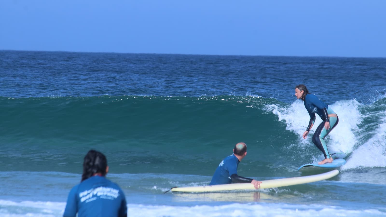 Ella riding a small breaking wave on a pale surfboard, crouched low, while two other people watch from the water nearby.
