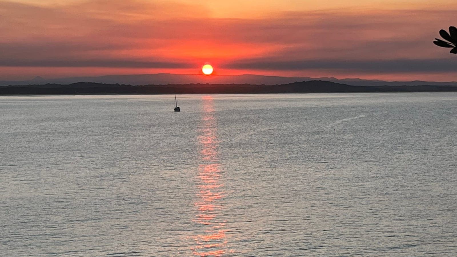 The sun sitting just above a low, dark headland at sunset, its light running in a long orange streak across calm water, with a single small yacht moored off to the left.