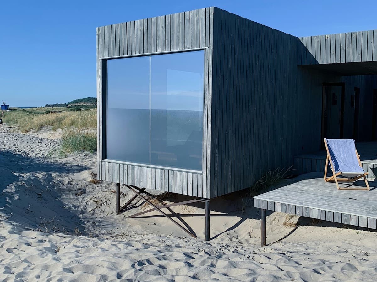 A timber-clad sauna cabin raised on legs above beach sand, with a striped deckchair on its decking under a clear blue sky.