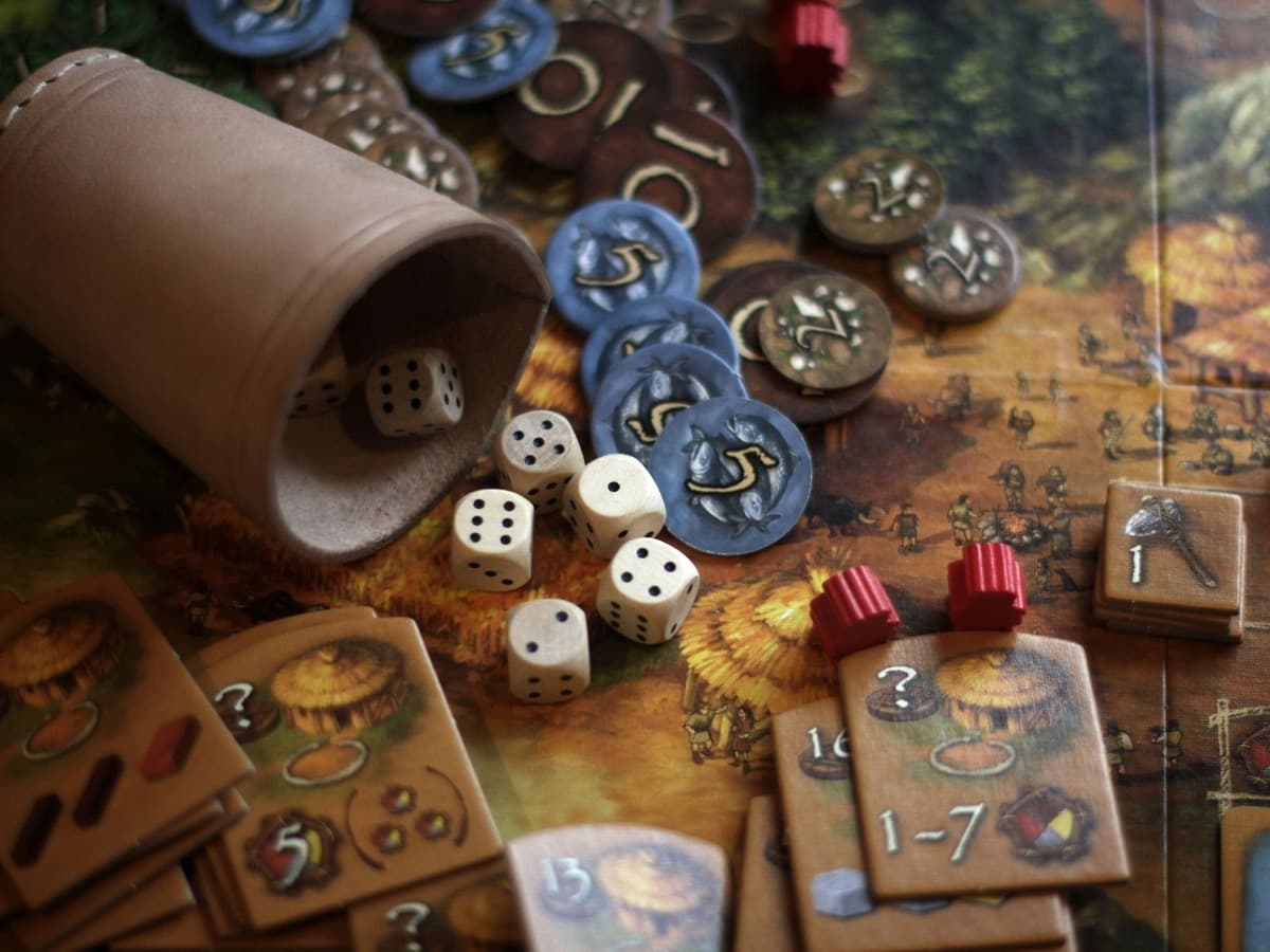 A toppled leather dice cup with scattered dice, wooden and blue tokens and playing cards spread across a board game.