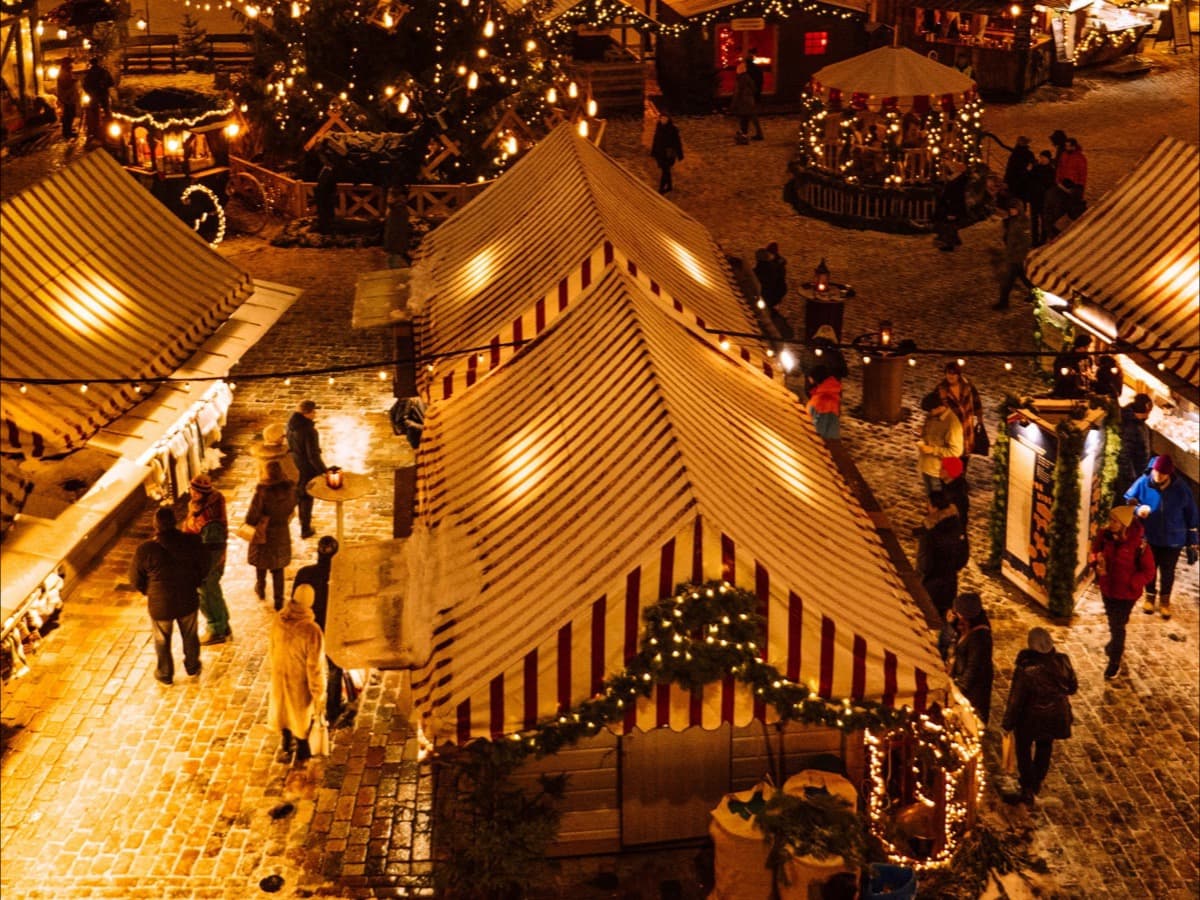 Rows of striped canvas market stalls lit with warm lights at night, seen from above, with people walking between them in the snow.