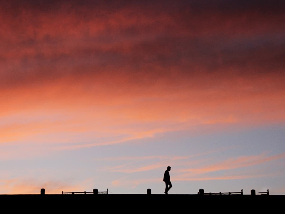 A lone figure walking along a raised path lined with low railings, silhouetted against a red and orange sunset sky.