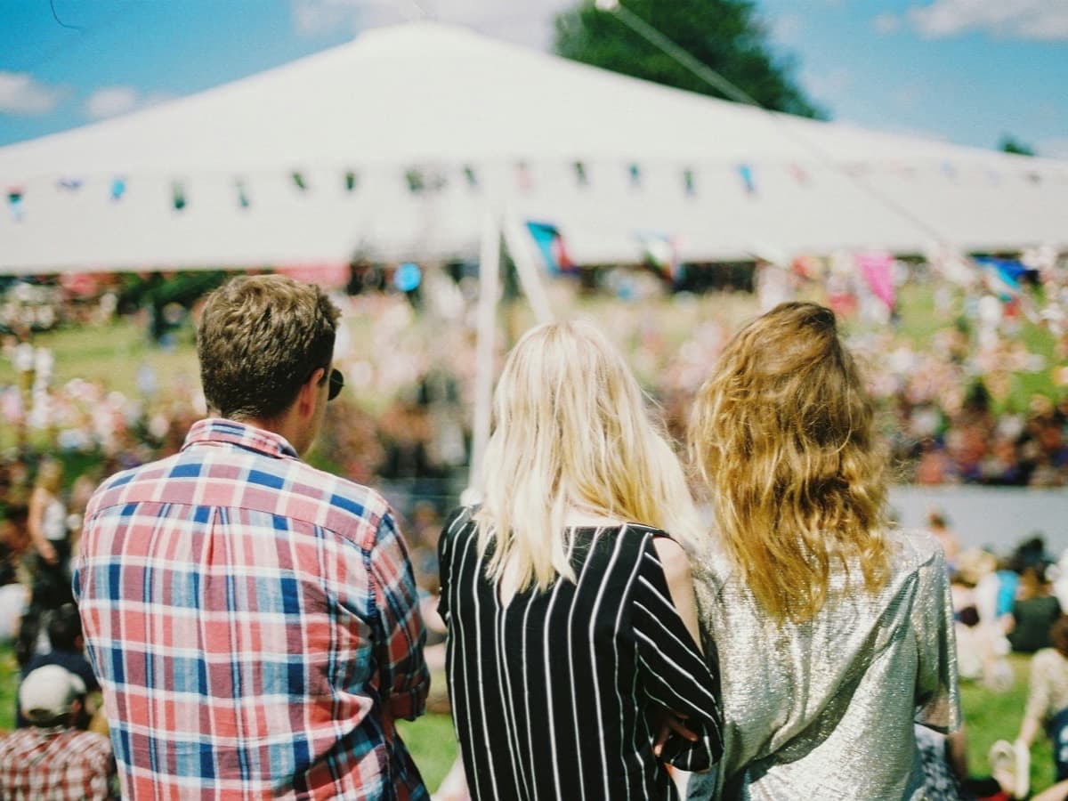 Three people seen from behind at an outdoor festival, looking across a grassy field towards a large white marquee strung with bunting.
