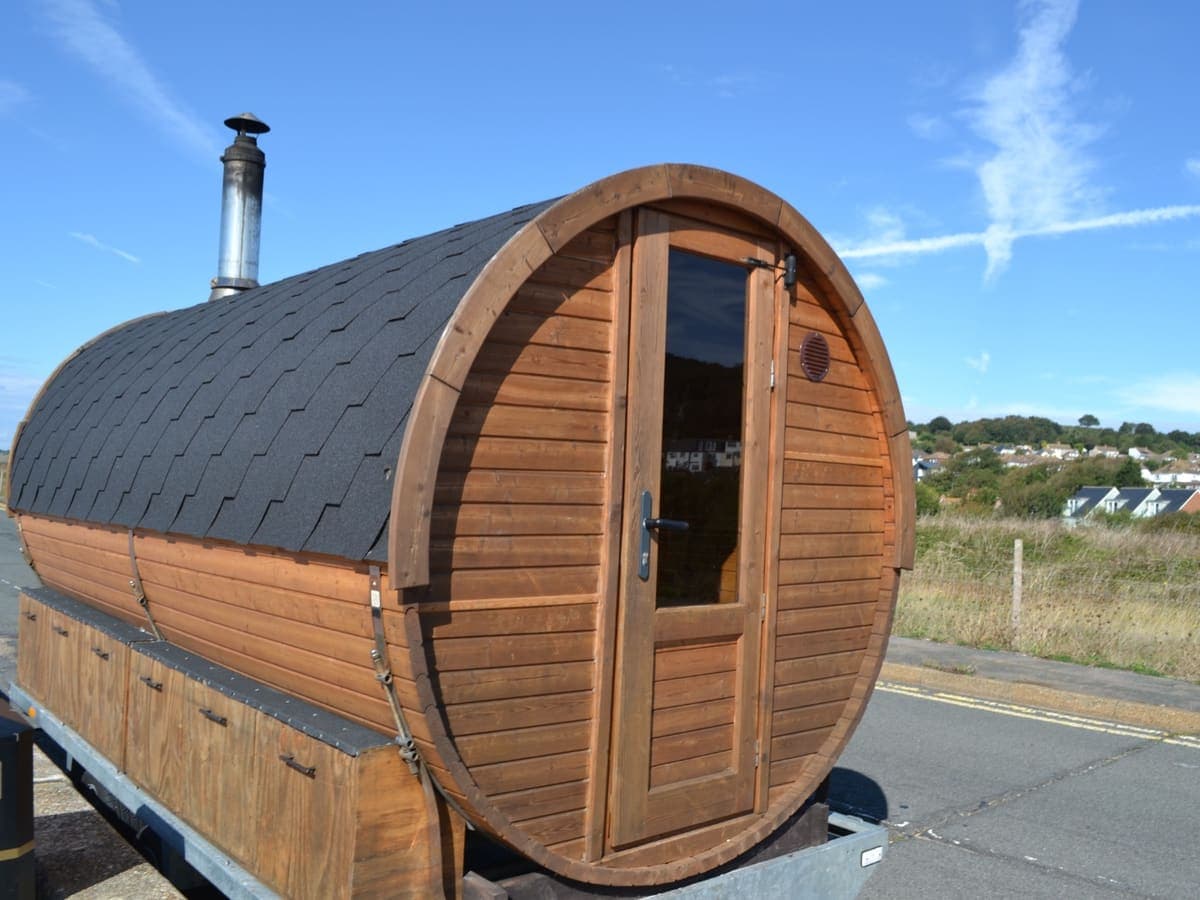 The Little Sauna, a wooden barrel sauna with a black shingle roof and a metal flue, on a trailer at the roadside under a blue sky, with houses on a green hillside behind.
