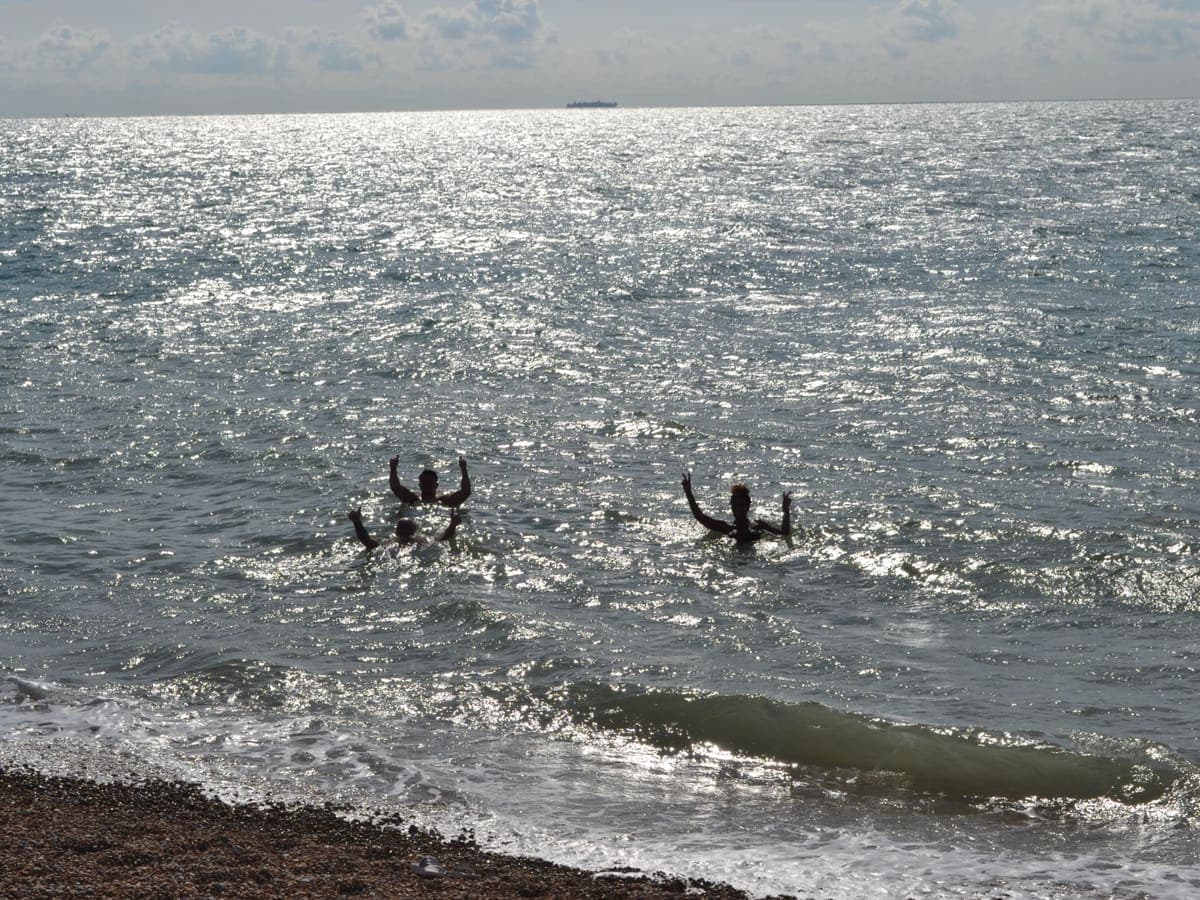 Three swimmers in silhouette with their arms raised in a sunlit, glittering sea, a shingle beach in the foreground and a ship on the horizon.
