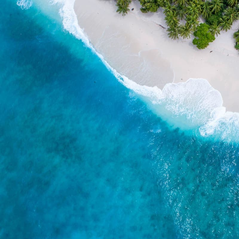 An aerial view of turquoise shallows meeting a pale sand beach, with surf breaking in a curve.