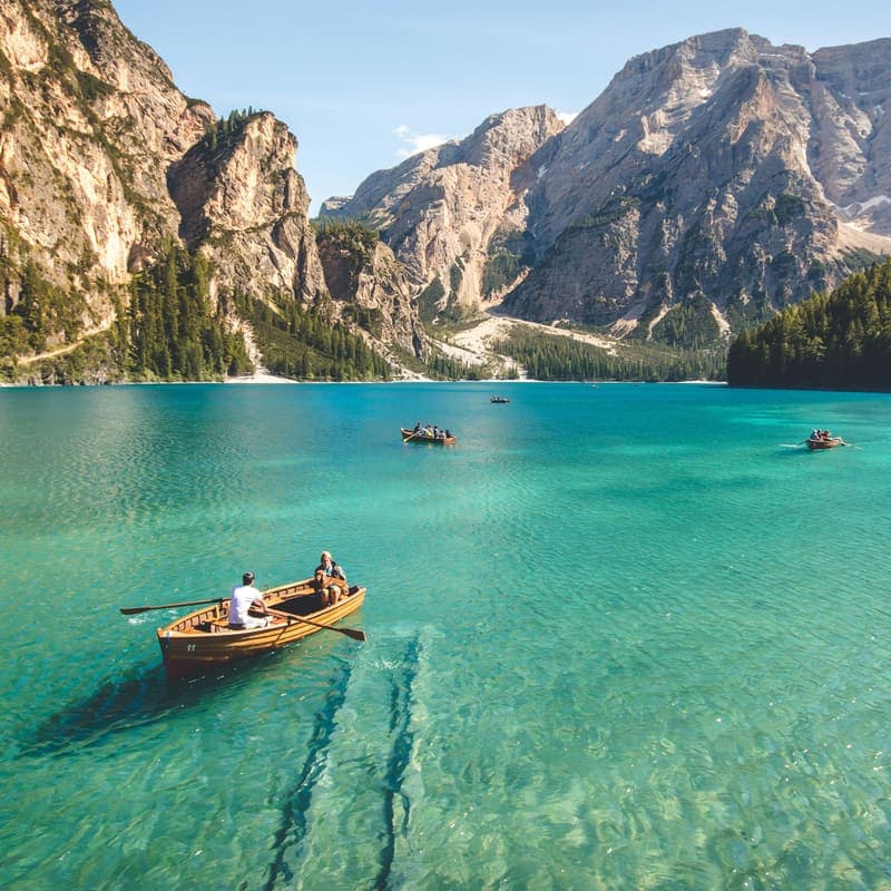 A small wooden rowing boat crossing a clear turquoise mountain lake, with steep pine slopes behind.
