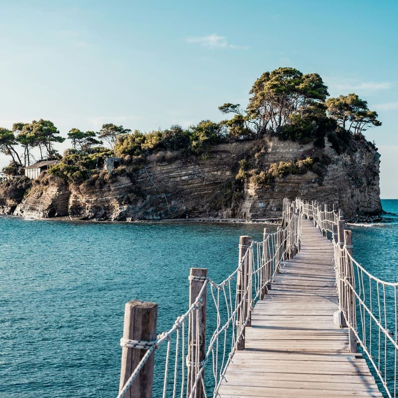 A wooden rope bridge running out to a tiny tree-covered island in blue-green water.