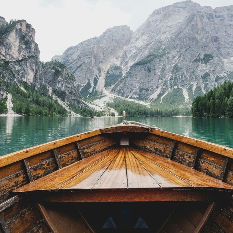 The varnished bow of a wooden boat on flat green water, facing a grey mountain face.