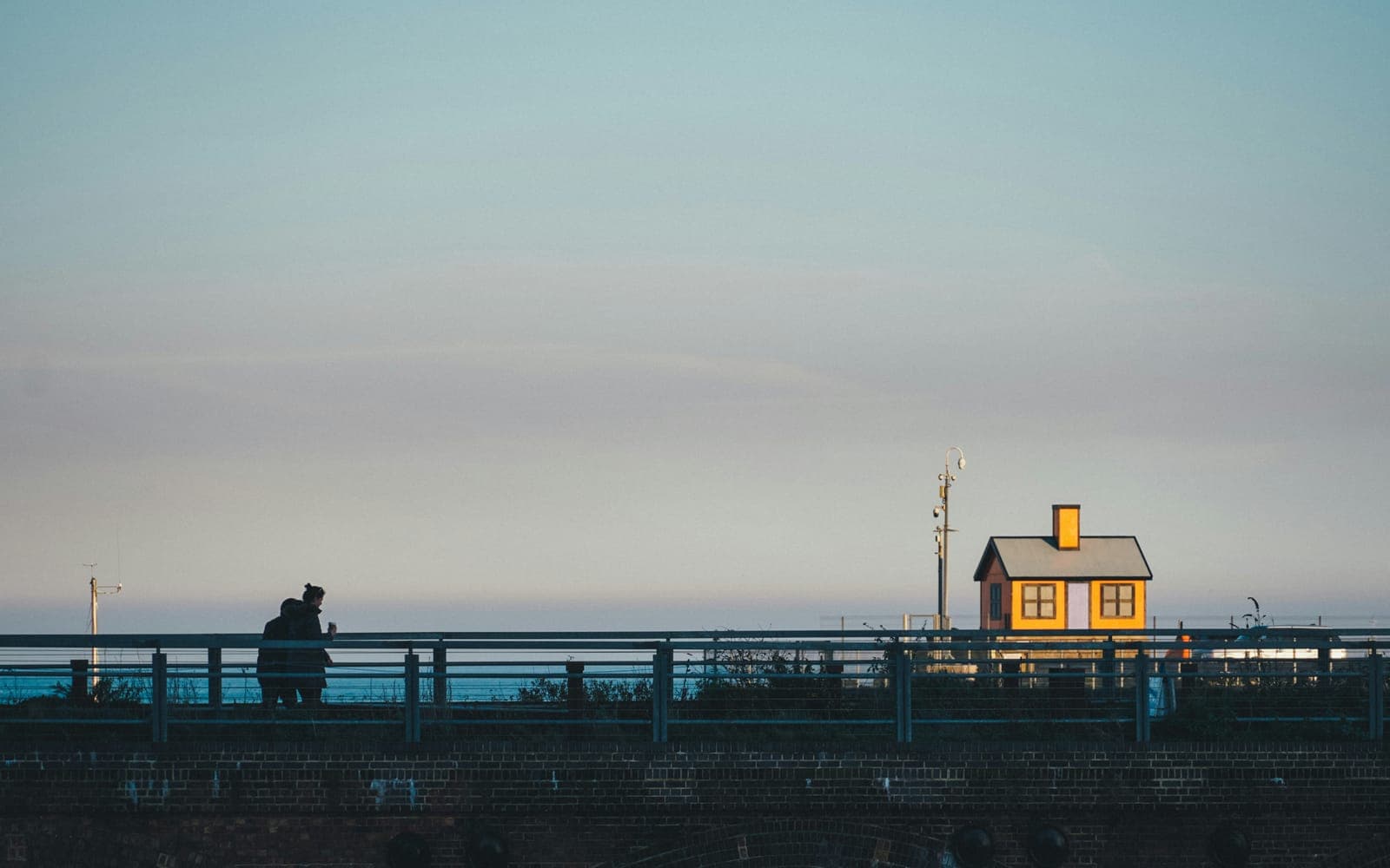 Two people standing together at a seafront railing at dusk, looking out over a flat sea towards a small lit hut.
