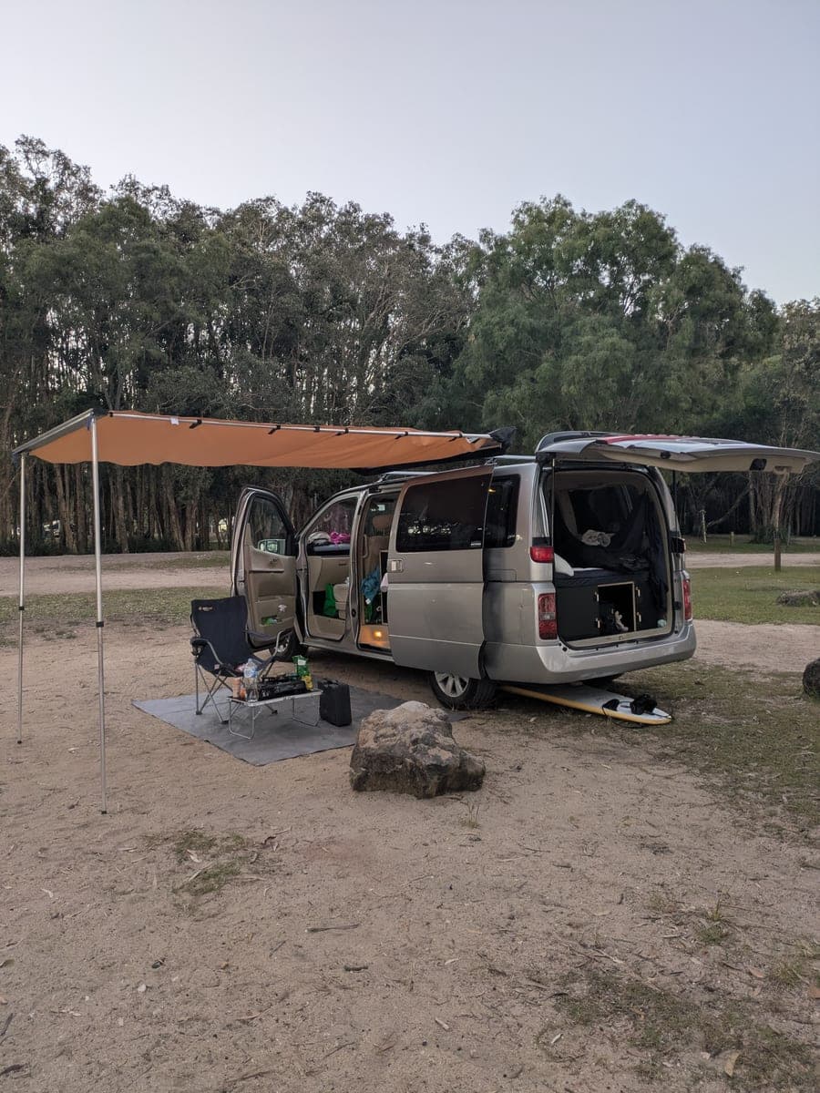 A small silver campervan parked on sandy ground at dusk with its side and rear doors open, an orange awning out over a camp chair and a low table, a surfboard lying underneath, and tall eucalyptus woodland behind.