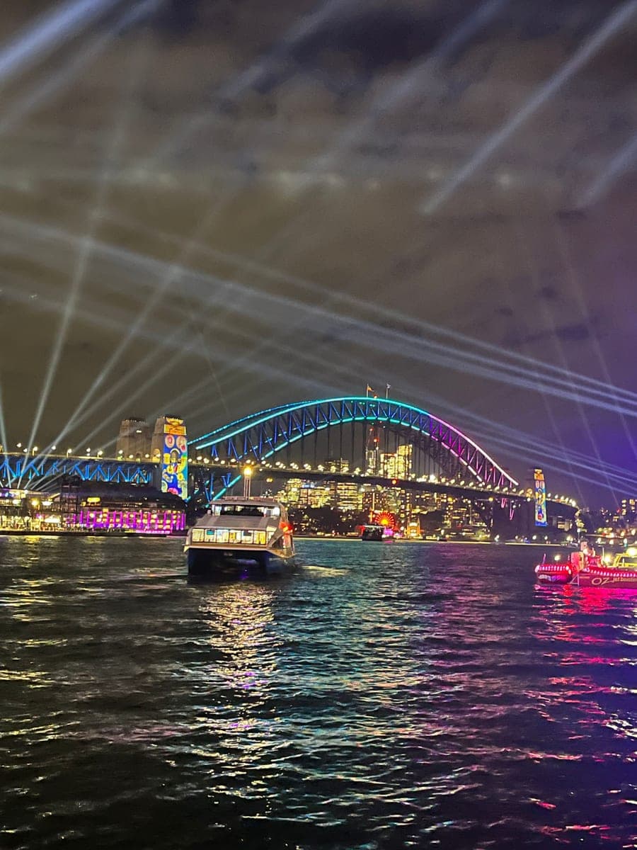 Sydney Harbour Bridge at night lit in turquoise and pink, searchlight beams fanning across the sky above it, with city lights and a passing ferry reflected in the water below.