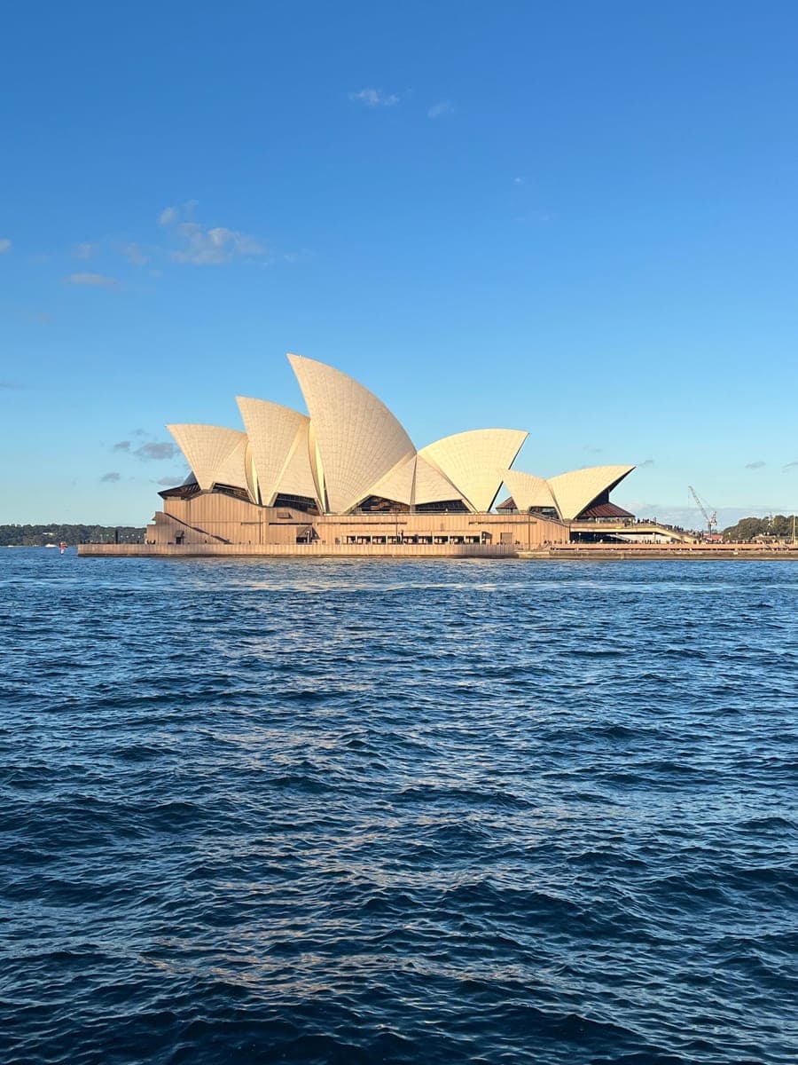 Sydney Opera House in late afternoon sun, its white shells side-on above the harbour, seen across choppy blue water.
