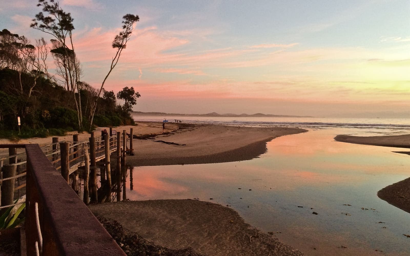 A sandy beach at sunset seen from beside a wooden boardwalk, with pink clouds reflected in a stream winding out across the sand towards the sea.
