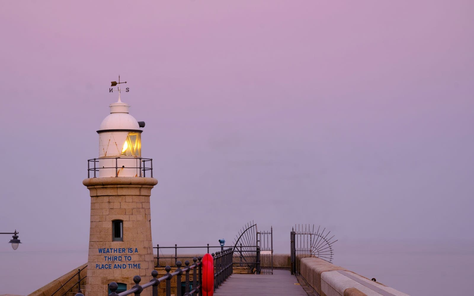 A harbour lighthouse at dusk under a soft pink sky, its lamp lit, with an empty walkway running out beside it.